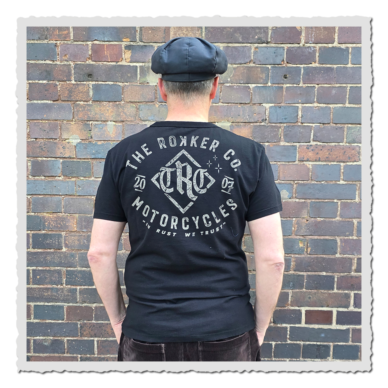 Rückansicht eines Mannes mit schwarzem T-Shirt, hellem Schriftaufdruck und Kappe vor rustikaler Ziegelwand.